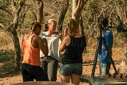 Four people are standing outside talking among trees in the sharp sun in Africa.