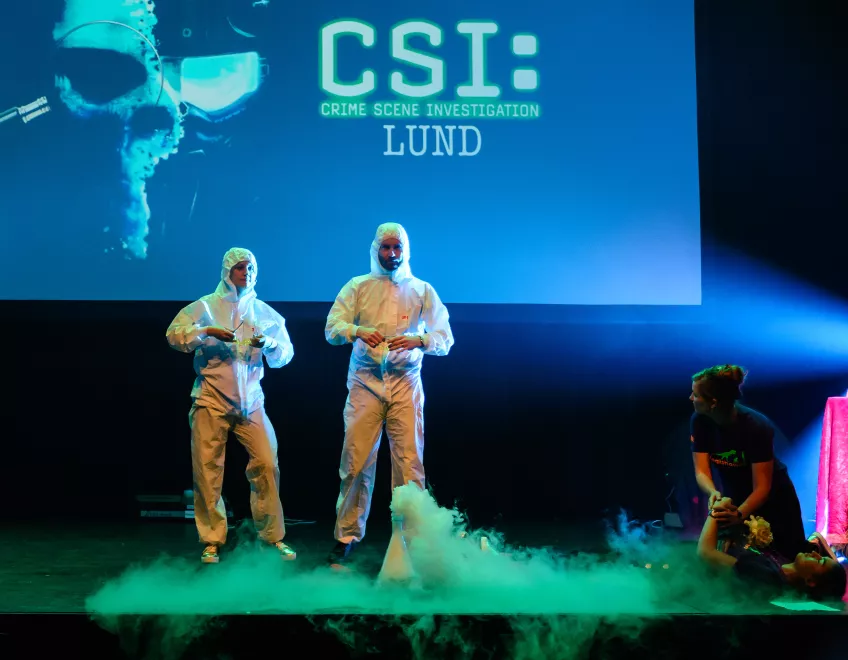 Two people are standing in white overalls on a stage where a green smoke is flowing. Photo.